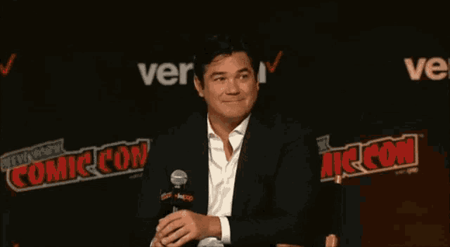 a man in a suit is sitting in front of a comic con sign