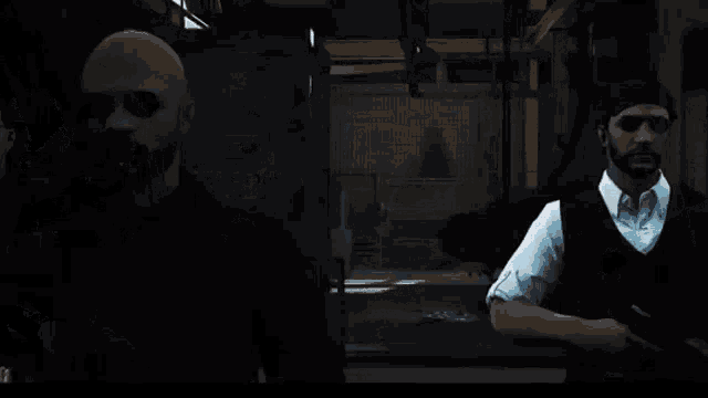 a man with a beard is standing next to another man with a gun in a dark room