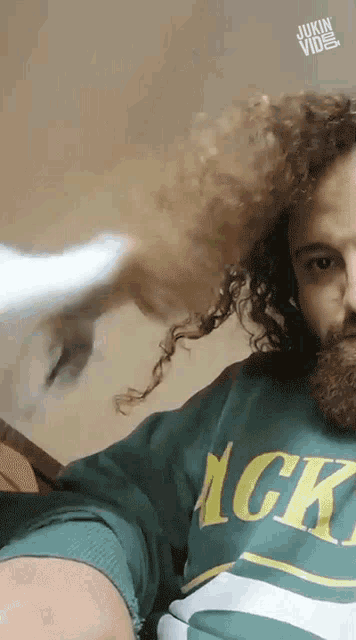 a man with curly hair and a beard wears a green shirt that says ' gkc ' on it