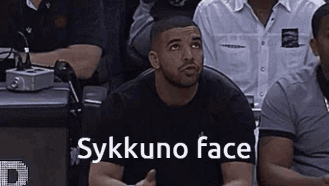 a man in a black shirt is sitting in a crowd with the words " sykkuno face " written on the bottom