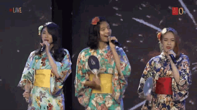 three women in kimonos singing into microphones in front of a screen that says live on it