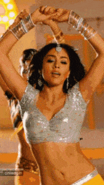 a woman in a crop top and yellow skirt is dancing with her hands in the air .