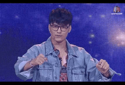 a young man wearing glasses and a denim jacket is standing in front of a blue screen .