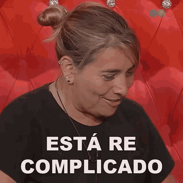 a woman is smiling in front of a red couch with the words esta re complicado written in white letters