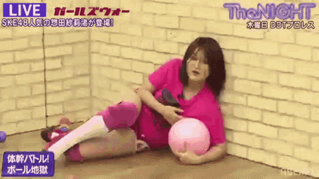 a woman in a pink shirt is laying on the floor holding a pink ball in front of a brick wall