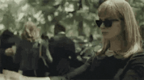 a woman wearing sunglasses is sitting at a table in a crowded park .
