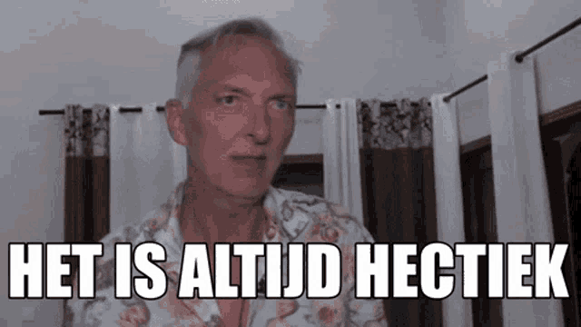 a man in a floral shirt is standing in front of a window with curtains and says het is altijd hectiek