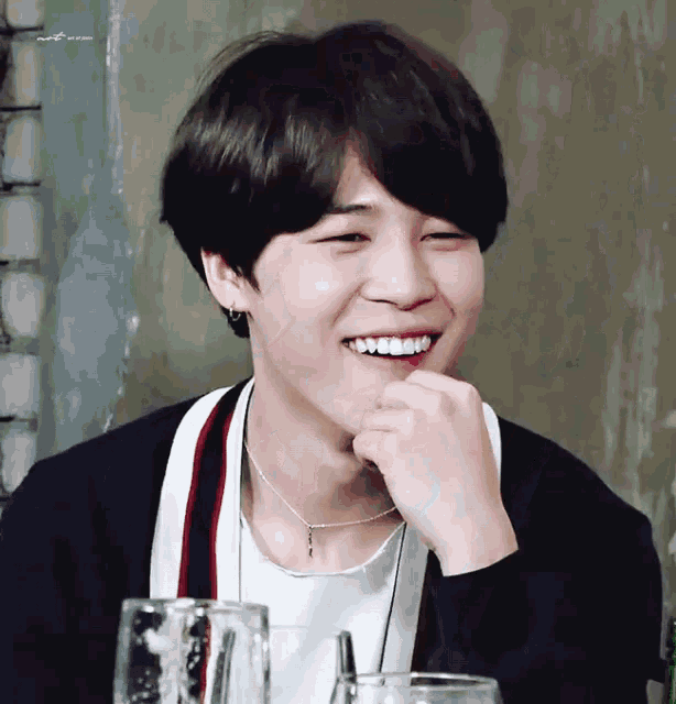 a young man wearing a necklace and a sweater smiles with his hand on his chin