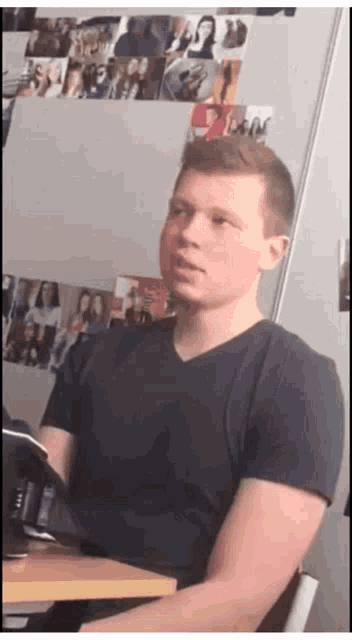a man in a black shirt is sitting in front of a wall with pictures of people on it