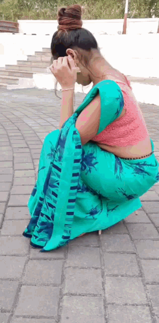 a woman in a blue saree and a pink blouse is squatting on a brick sidewalk .