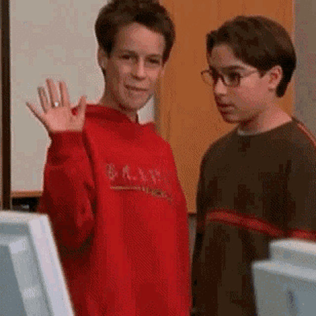 two young boys are standing next to each other in a room and one of them is waving at the camera .