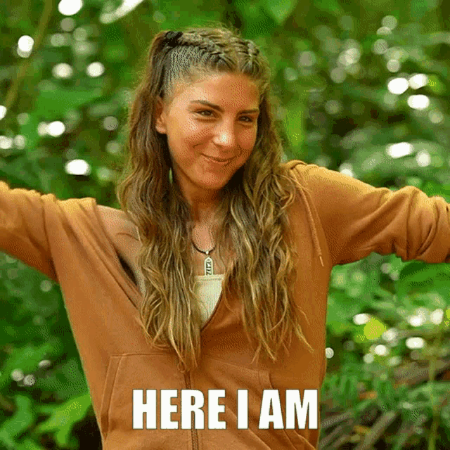 a woman in a brown hoodie is standing in the woods with her arms outstretched and the words here i am below her .
