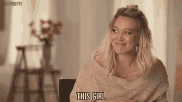 a woman in a sweater is sitting in a chair and smiling while saying this girl .
