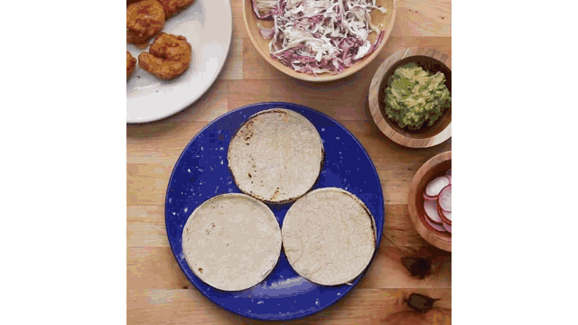three tortillas on a blue plate next to a bowl of guacamole and a bowl of coleslaw