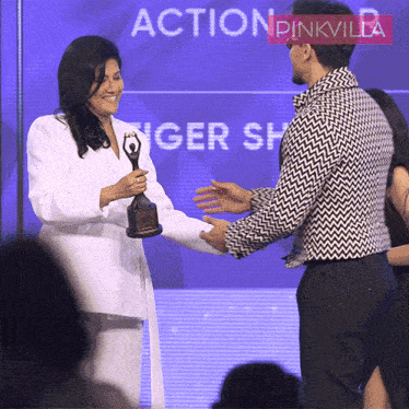 a woman in a white jacket is holding a trophy and shaking hands with a man in a black and white jacket