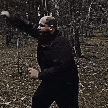 a man in a black jacket is standing in the woods holding a stick .