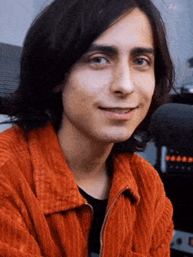 a young man with long black hair is wearing an orange jacket and smiling .