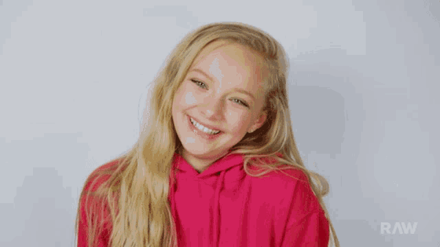 a girl in a pink hoodie is smiling in front of a white background with raw written on it