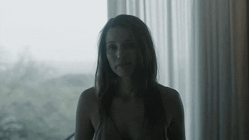 a woman in a tank top is standing in front of a window looking out .