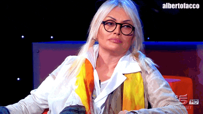 a woman wearing glasses and a scarf sits in front of a sign that says albertofacco