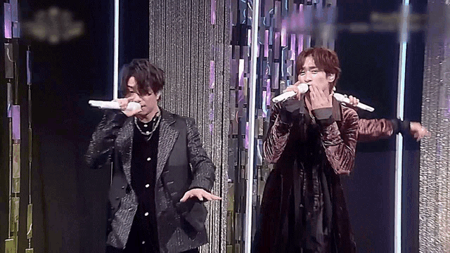 two men singing into microphones on a stage with a curtain behind them