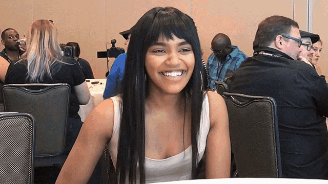 a woman in a white tank top smiles while sitting at a table in a room with other people