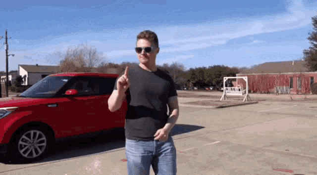 a man wearing sunglasses and a black shirt stands in front of a red car in a parking lot