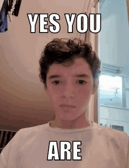 a young boy with curly hair is wearing a white t-shirt and a meme .