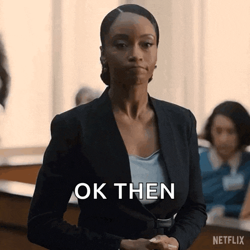 a woman in a suit is standing in a courtroom with her hands folded and says ok then .
