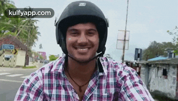 a man wearing a helmet and a plaid shirt is smiling while riding a motorcycle down a street .