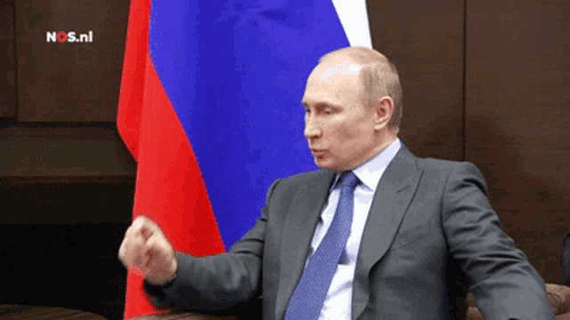 a man in a suit and tie sitting in front of a russian flag with nos.nl written on the bottom