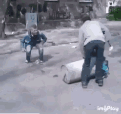 a man is squatting down next to a barrel while another man stands behind him .