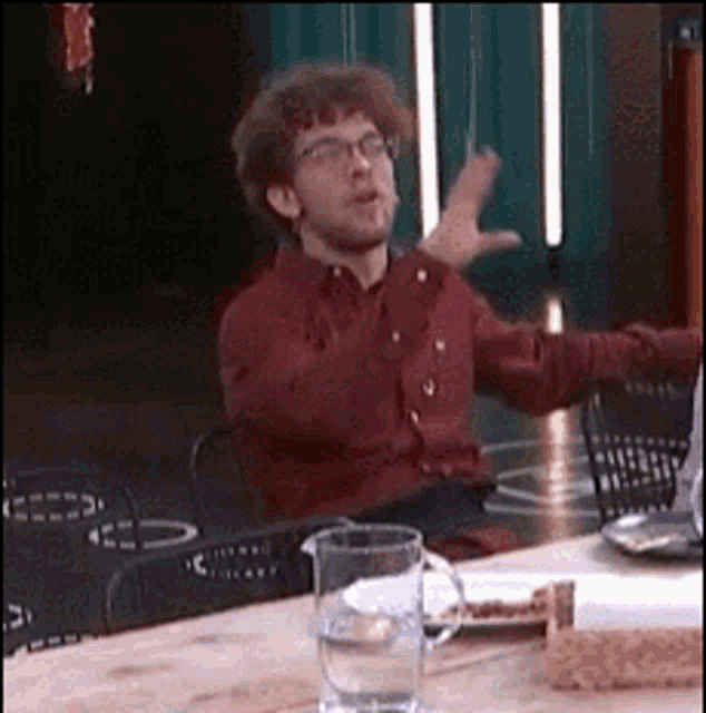 a man in a red shirt and glasses is sitting at a table with his arms outstretched