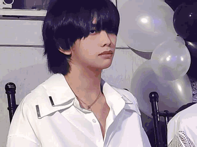 a man wearing a white shirt and a necklace is sitting in front of balloons