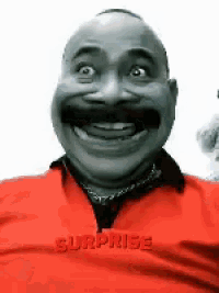 a bald man with a mustache is smiling and wearing a red shirt with surprise written on it
