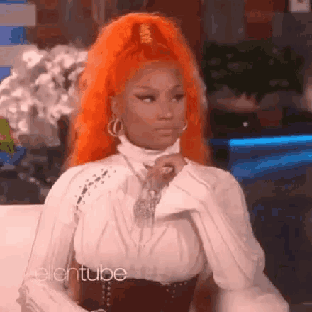 a woman with orange hair is sitting on a couch wearing a white shirt and earrings .