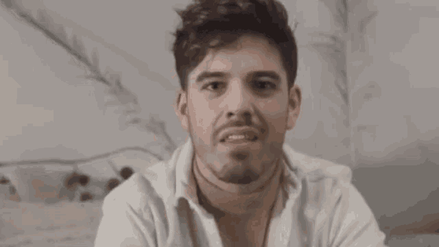 a man with a beard and a white shirt is sitting on a couch and looking at the camera .