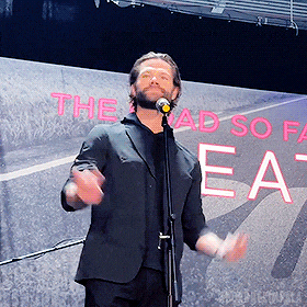 a man is singing into a microphone in front of a sign that says " the road so far "