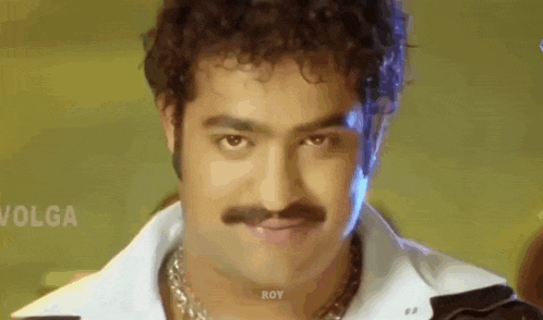 a man with curly hair and a mustache is wearing a white shirt and black jacket .