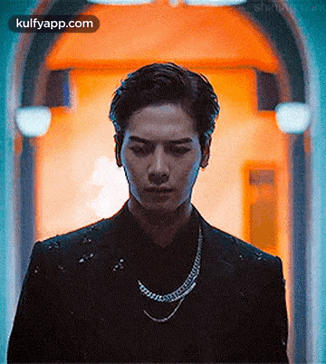 a man wearing a black suit and a necklace is standing in front of a door .
