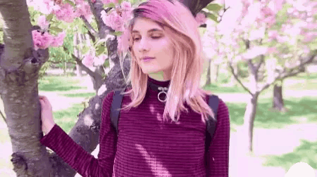 a woman with pink hair is standing next to a tree in a park with flowers .