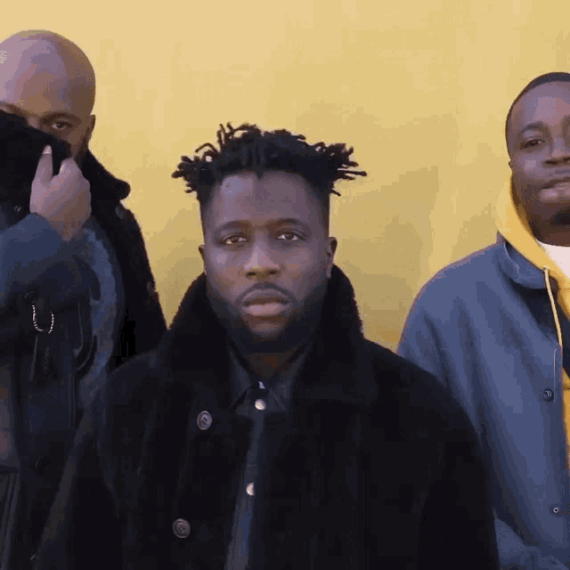 three men are standing in front of a yellow wall and one has a beard