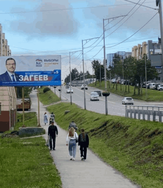 a group of people walking down a sidewalk in front of a billboard for a political party