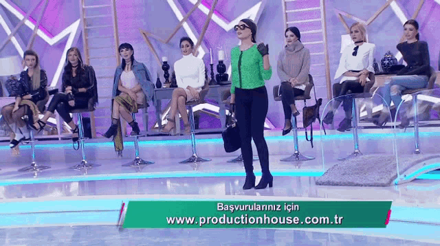 a woman in a green jacket stands in front of a group of women sitting on stools