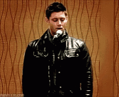 a man in a leather jacket is singing into a microphone in front of a brown wall