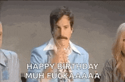 a man with a mustache is sitting in front of a group of people and says `` happy birthday muh fuck aaa '' .