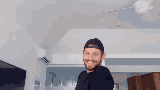 a man with a beard wearing a black shirt and a baseball cap is smiling in front of a ceiling fan