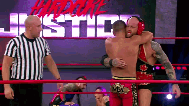 two men are hugging in a wrestling ring with a referee standing behind them .