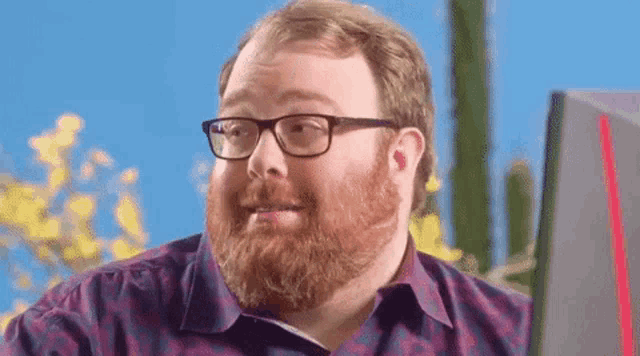 a man with a beard wearing glasses is sitting in front of a computer monitor .