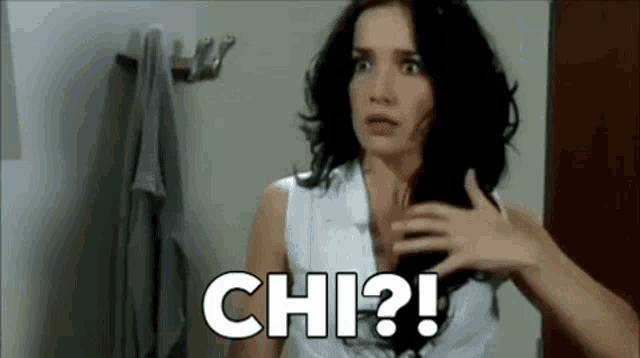 a woman in a white shirt is holding her hair in a bathroom and says chi ?